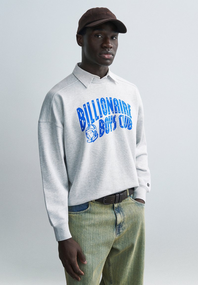 Γκρι φούτερ με μπλε γραφικό «Billionaire Boys Club», πουκάμισο με γιακά από κάτω, συνδυασμένο με ανοιχτόχρωμο τζιν. Φοράει καφέ καπέλο.