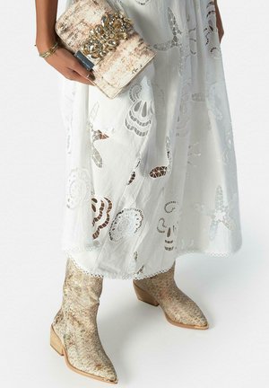 Mujer con un vestido blanco bordado, sosteniendo un bolso de mano adornado en dorado y llevando botas con punta afilada de piel de serpiente dorada y tacones gruesos.