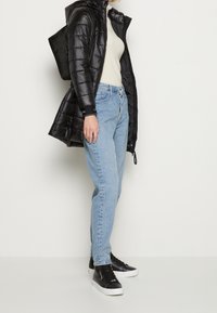Donna che indossa una giacca a vento nera, un maglione color crema, jeans azzurri chiari, sneaker alte nere e porta uno zaino nero su uno sfondo bianco.