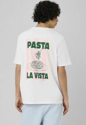 Personne aux cheveux bouclés foncés de profil, portant un t-shirt blanc à rayures roses avec le texte vert "PASTA LA VISTA" et une illustration d'une assiette de spaghettis.