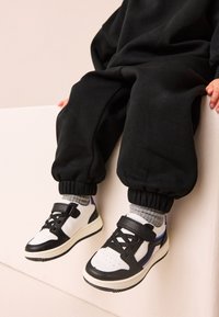 Child wearing black pants and gray socks sitting on a white ledge, showing white sneakers with black and purple accents and Velcro straps.
