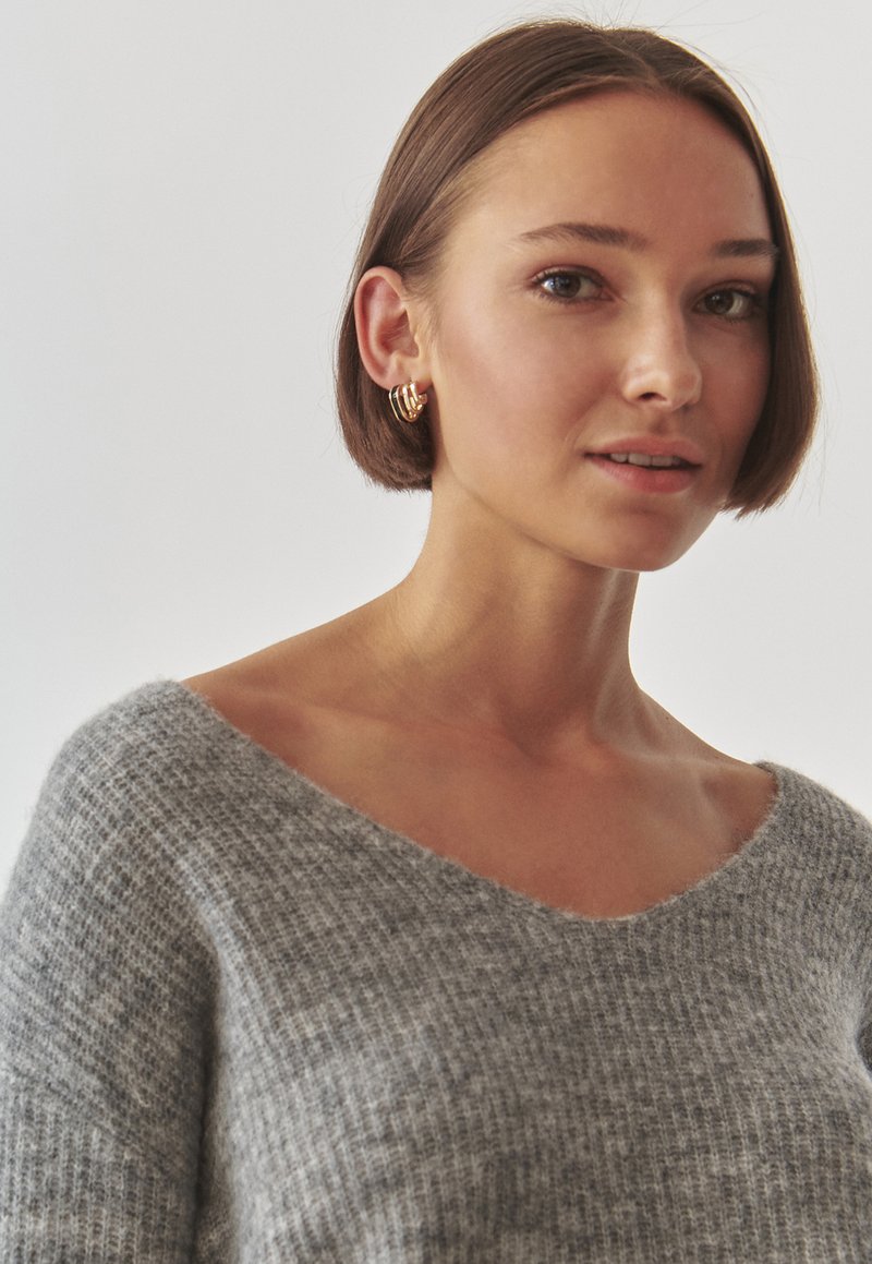 Szary, dziany sweter z szerokim dekoltem w kształcie litery V i prążkowanym teksturowaniem. Złote kolczyki w kształcie obręczy podkreślają stylizację. Miękka tkanina, luźny krój.