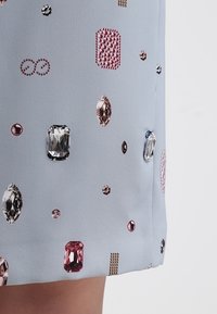 Jasnoniebieska tkanina z różnymi teksturowymi zdobieniami, w tym kryształami w różnych kształtach i kolorach, rozmieszczonymi po powierzchni.