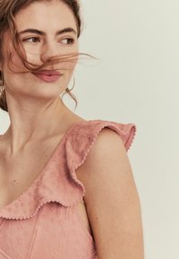 Rosa ärmelloses Kleid aus texturiertem Stoff, mit rüschendetails an den Schultern und einem kleinen gewellten Saum; das Modell hat lockere, wellige Haare.