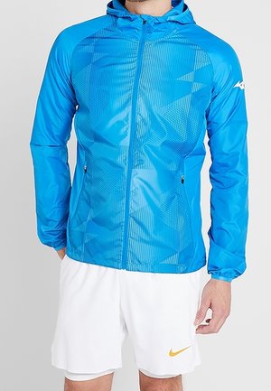 Homme portant une veste à capuche bleu vif avec des motifs géométriques et un short blanc avec un petit logo jaune Nike sur la jambe gauche.