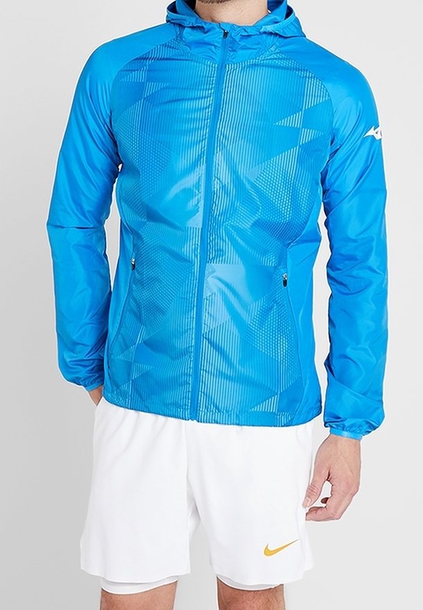Homme portant une veste à capuche bleu vif avec des motifs géométriques et un short blanc avec un petit logo jaune Nike sur la jambe gauche.