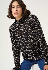 Schwarze Bluse mit abstraktem beigem Muster, hohem Kragen und langen Ärmeln, aus einem strukturierten Stoff. Getragen mit blauen Jeans.