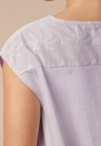 Haut lilas clair avec des motifs floraux brodés le long de l'encolure et des épaules. Manches courtes, texture douce et coupe décontractée.