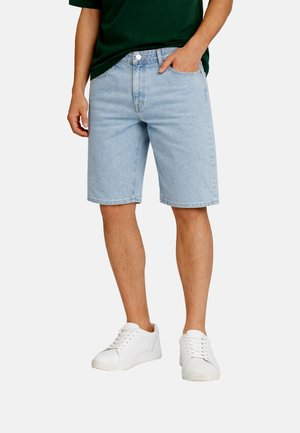 Mann kledd i lyseblå denimshorts, mørkegrønn skjorte og hvite joggesko, stående med en hånd i lomma.