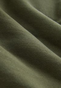 Oliijfgroene stof met een gladde textuur en lichte rek, met een subtiel gebreid patroon, gevouwen om diepte en contour te tonen.