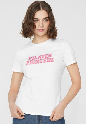 Mladá žena s krátkými hnědými vlasy, která má na sobě bílé tričko s růžským textem "PILATES PRINCESS", drží lem trička, na jednobarevném pozadí.