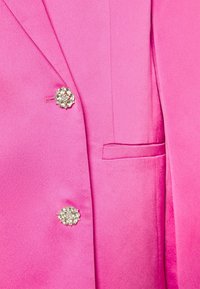 Blazer rose vif avec deux boutons ornés en forme de fleur en cristal et une petite poche fendue sur le côté droit.