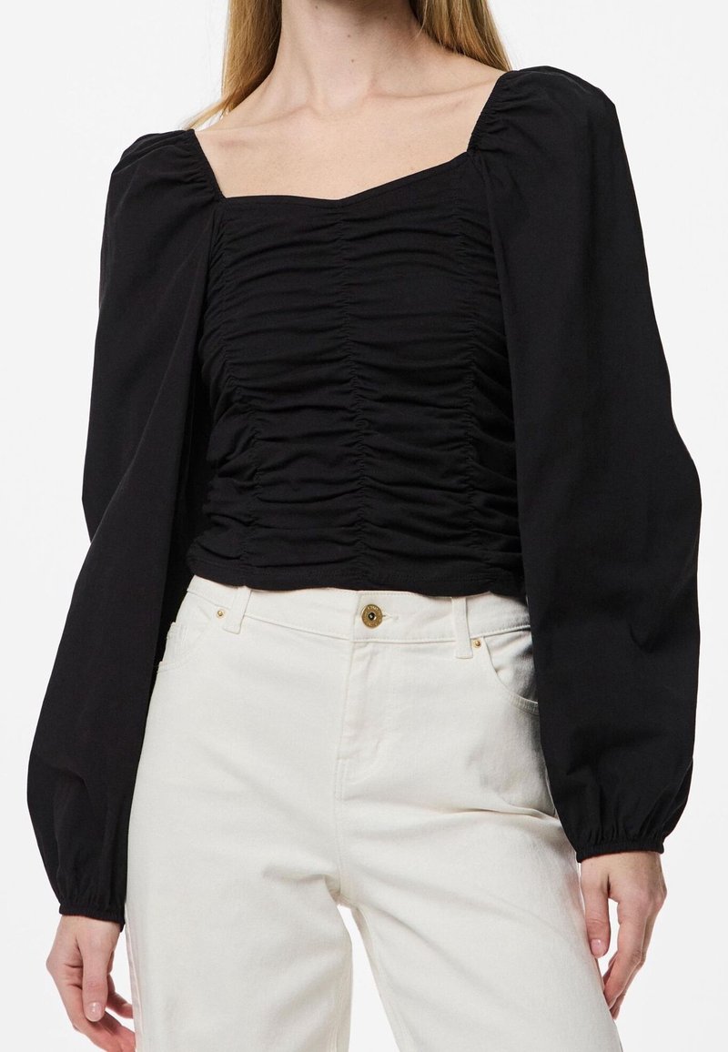 Femme portant un chemisier noir froncé à col carré avec des manches longues bouffantes et un pantalon blanc taille haute à patte de boutonnage.