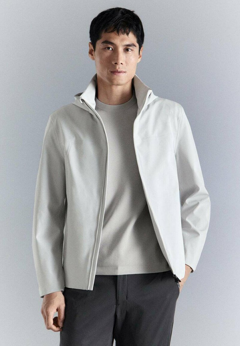 Homme portant une veste zippée gris clair sur une chemise assortie, debout avec une main dans la poche devant un fond gris uni.
