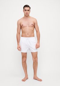 Pantaloni scurți de înot albi, cu talie ajustabilă, având un detaliu discret cu logo. Materialul este neted și ușor. Modelul stă desculț.