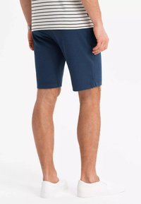 Marineblaue Shorts aus weichem Stoff, mit gerader Schnittführung und seitlichen Schlitzen für Bewegungsfreiheit. Kombiniert mit weißen Schlupfschuhen.