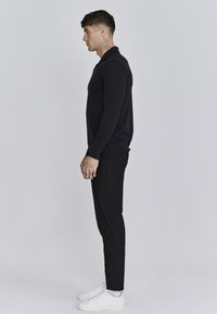 Polo de manga larga negro, ajuste slim, combinado con pantalones negros y zapatillas blancas. Textura suave, diseño minimalista y sin patrones visibles.