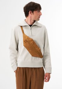 Braune Nylon-Crossbody-Tasche mit flachem Design, ausgestattet mit einem Reißverschluss und zwei runden Akzenten, getragen über einem hellgrauen Pullover.