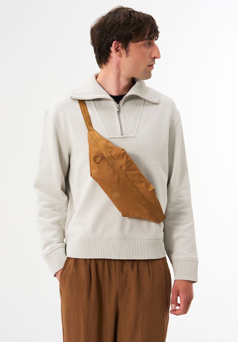 Braune Nylon-Crossbody-Tasche mit flachem Design, ausgestattet mit einem Reißverschluss und zwei runden Akzenten, getragen über einem hellgrauen Pullover.