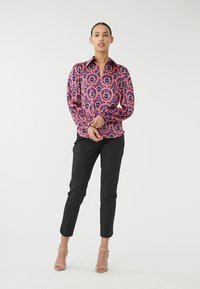 Kleurrijke blouse met een patroon van cirkelvormige vormen in roze en paars, gecombineerd met zwarte broek. De blouse heeft een kraag en lange mouwen.