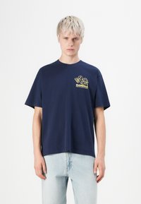 Blond person wearing navy blue T-shirt with yellow cartoon turtle and “KAMERA electronics store” text, paired with light blue jeans.