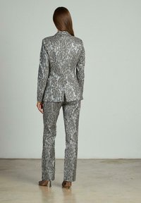 Completo jacquard argento con motivi floreali. La giacca aderente presenta uno spacco, mentre i pantaloni sono sartoriali con un taglio dritto, entrambi con una finitura lucida.