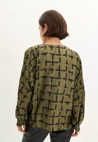 Blusa verde oliva ampia con un motivo geometrico nero, maniche lunghe con polsini arricciati e collo rotondo.