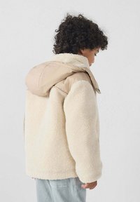 Chaqueta de borreguito cremosa y suave con capucha de nylon beige clara, que presenta un ajuste relajado y una superficie texturizada.