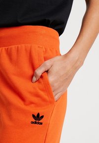 Oranžové bavlněné jogger kalhoty s pružným pasem na šňůrku, bočními kapsami a černým logem Adidas na levém stehně. Hladký materiál a pohodlné nošení.