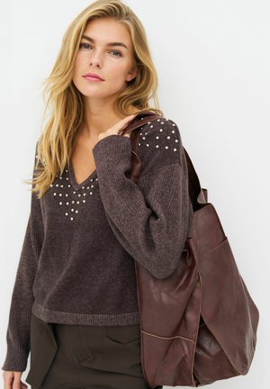 Femme blonde portant un pull en maille marron avec des détails en perles sur les épaules et la poitrine, portant un grand sac cabas en cuir marron sur une épaule.