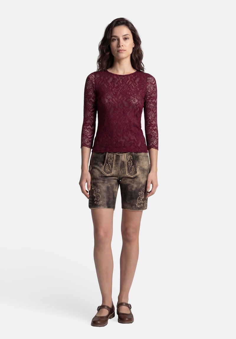 Frau steht und trägt ein bordeauxfarbenes Spitzenoberteil mit dreiviertellangen Ärmeln, braune bestickte Leder-Shorts und braune Mary-Jane-Schuhe.