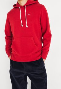 Homme portant un sweat à capuche rouge Levi’s avec des cordons blancs et un pantalon en velours côtelé noir, les mains dans les poches, sur un fond blanc.