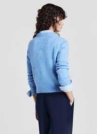Pull en tricot bleu clair avec une finition texturée, ourlet côtelé et poignets retroussés, porté sur une chemise rayée blanche et bleue, avec un pantalon foncé.