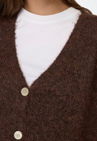 Gros plan d'un cardigan en maille marron duveteux avec deux boutons blancs sur un t-shirt blanc uni à col rond.