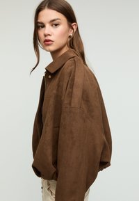 Giacca oversize in camoscio marrone con colletto classico, chiusura frontale con zip e grande tasca applicata, caratterizzata da una texture morbida e una vestibilità comoda.