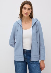 Felpa con zip di colore azzurro chiaro realizzata in tessuto testurizzato, con tasca frontale, cappuccio regolabile con cordino e polsini a coste. Indossata sopra un top bianco.