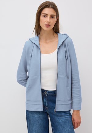 Lichtblauwe zip-up hoodie van textuurstof, met een voekzak, verstelbare trekkoorden in de capuchon en ribgebreide manchetten. G gedragen over een witte top.