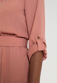 Manga arremangada y abotonada en una blusa texturizada de rosa polvo, metida en pantalones de cintura alta a juego con un cinturón de tela.