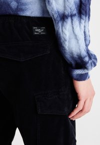 Czarne sztruksowe spodnie cargo z bocznymi kieszeniami, z brandowaną metką z tyłu. Niebieski sweter w stylu tie-dye z teksturowanymi dzianinowymi rękawami.