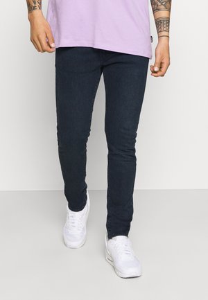 Personne portant un jean noir slim, des baskets blanches et un t-shirt lavande, marchant devant un fond clair uni.