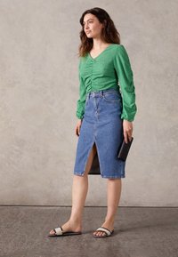 Donna che indossa un top verde a maniche lunghe arricciato, una gonna di jeans blu a lunghezza ginocchio con spacco frontale, tiene una pochette nera e sandali beige.