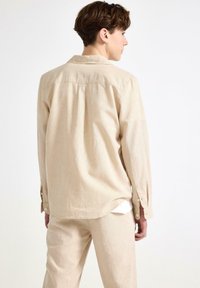 Camicia lunga beige chiaro con taglio dritto, polsini a bottone e dettagli con pieghe sul retro. Tessuto morbido e leggero, abbinata a pantaloni coordinati.