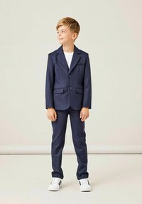 Costume bleu marine avec une veste à simple bouton et deux poches avant, associé à un pantalon assorti. Porté par-dessus un t-shirt blanc et des baskets blanches.