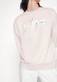 Personne portant un sweat-shirt rose clair avec un logo multicolore "Hugo 1985" sur la poitrine et un pantalon blanc.