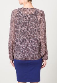 Blouse à manches longues en tissu transparent, bleu marine avec imprimé abstrait rose et blanc ; ourlet arrondi et poignets élastiques. Portée avec une jupe bleue unie.