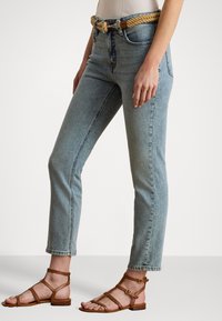 Ljusblå denimjeans har hög midja och smala ben, kompletterade av ett flätat bälte i tan-färg. Bruna sandaler med remmar fulländar outfiten.