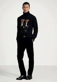 Camisola de gola alta em azul-marinho escuro com dois motivos de ursinhos vestidos de fato formal, combinada com calças pretas e botins pretos. Textura de malha macia.