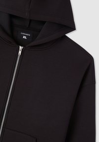 Felpa nera con zip realizzata in tessuto liscio, dotata di tasca frontale, un cappuccio grande e una zip argentata. L'etichetta mostra la taglia XL.
