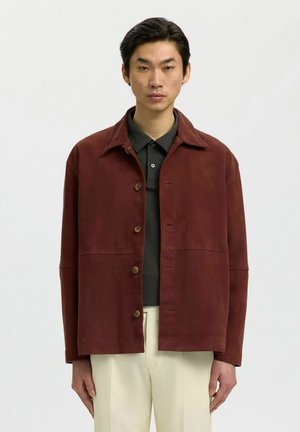 Homme portant une veste marron boutonnée sur un polo foncé et un pantalon beige clair, debout devant un fond blanc uni.
