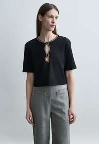 Mujer con un top negro de manga corta con adornos de cuentas doradas en el escote y un recorte, combinado con pantalones de talle alto grises y con textura.